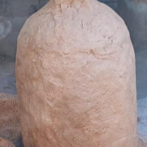 May include: A tall, cylindrical clay jug with a narrow neck and a textured, uneven surface in a light brown earth tone. The vessel features a hand-molded appearance with visible ridges and a small logo tag reading KVK Designs.