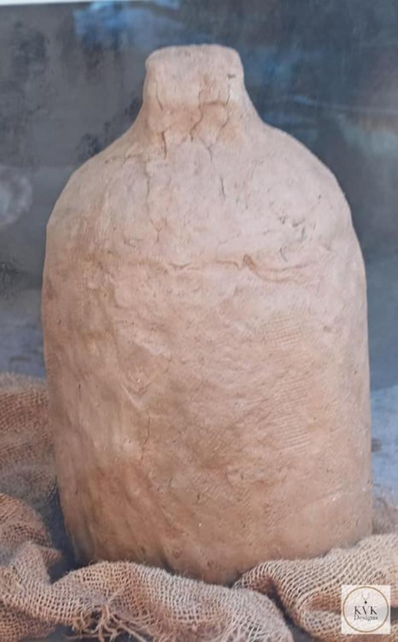 May include: A tall, cylindrical clay jug with a narrow neck and a textured, uneven surface in a light brown earth tone. The vessel features a hand-molded appearance with visible ridges and a small logo tag reading KVK Designs.