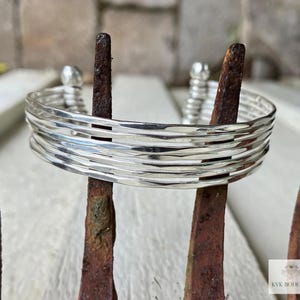 May include: A silver bracelet made of multiple thin bands, each with a hammered texture. The bracelet is stacked on top of itself, creating a layered effect.