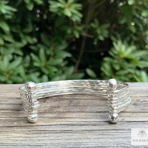 May include: A silver bangle bracelet with a hammered finish and rounded ends. The bracelet is made of multiple strands of wire that are woven together.