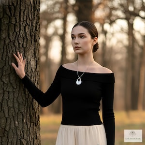 May include: A woman wearing a black off-the-shoulder long-sleeve top and a cream-colored skirt. A silver pendant necklace hangs around her neck. The background is a blurred forest scene.
