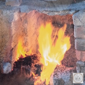 May include: A bright orange and yellow fire burns within a rustic stone and brick fireplace. Glowing embers and small sparks are visible amidst the flames. A small white square logo with the text KVK Designs is in the bottom corner.