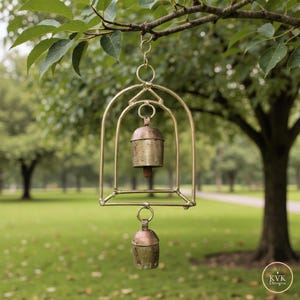 May include: A brass wind chime with two bells suspended within a decorative frame. The chime hangs from a tree branch against a blurred green background. The bells have a rustic, aged appearance.