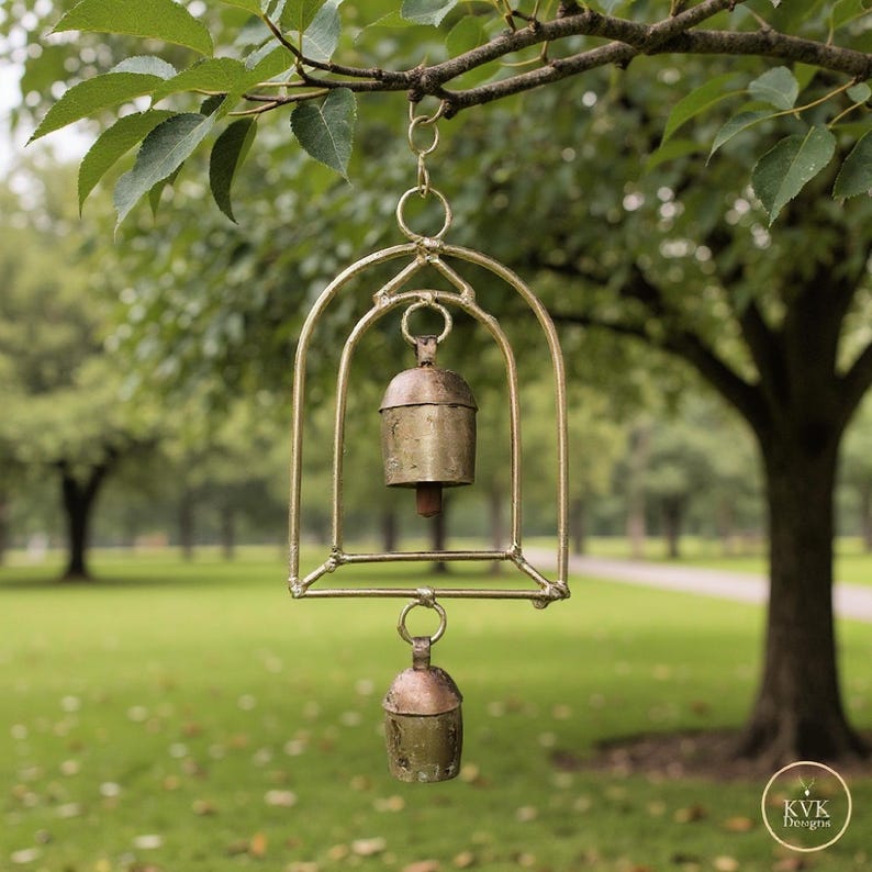 May include: A brass wind chime with two bells suspended within a decorative frame. The chime hangs from a tree branch against a blurred green background. The bells have a rustic, aged appearance.