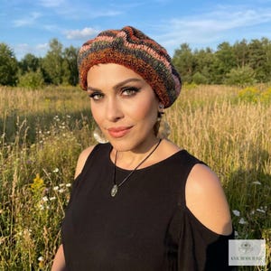 May include: A knit beret featuring a multi-colored horizontal striped pattern in shades of orange, brown, green, and black. The hat has a textured, soft appearance and is worn on the head. A small white logo with the text KVK BOHOFAIR is visible.