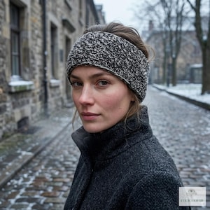May include: A woman wearing a black and white knit headband and a dark gray coat. The headband is thick and covers the forehead and ears. The background is a cobblestone street with buildings.
