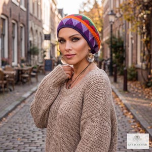 May include: A multi-colored crochet beanie featuring a geometric triangle pattern in shades of purple, orange, green, and red. The beanie is worn with a beige chunky knit sweater and silver circular drop earrings. The text KVK BOHOFAIR is visible.