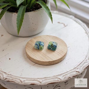 May include: Two square stud earrings with a mosaic design in shades of blue and green. The earrings are displayed on a small, round, light-colored wooden coaster. A potted plant is in the background.