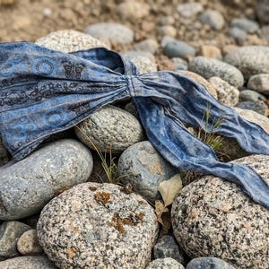 May include: A blue patterned headband with a decorative design, lying on a bed of gray rocks. The headband features a repeating pattern of circles and floral elements. The fabric has a faded, vintage appearance.