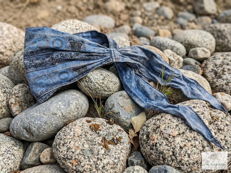 May include: A blue patterned headband with a decorative design, lying on a bed of gray rocks. The headband features a repeating pattern of circles and floral elements. The fabric has a faded, vintage appearance.