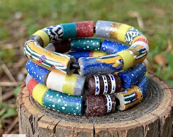 May include: Three colorful beaded bracelets stacked on a wooden surface. The bracelets feature a variety of colors and patterns, including yellow, blue, red, and white. The beads are cylindrical and strung together with a black cord.