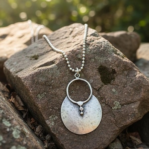 May include: A silver-toned pendant necklace with a large, textured circular pendant and a smaller, round pendant. The necklace is displayed on a rough, brown rock, with a blurred green background. The necklace has a ball chain.