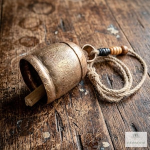 May include: A brass bell with an etched floral and geometric pattern, featuring a wooden clapper inside. A jute rope is attached to the top loop, decorated with a black bead and three light brown wooden beads. The bell has a weathered finish.