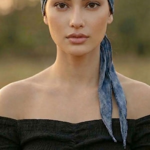 May include: A woman wearing a blue patterned headband and a black off-the-shoulder top. The headband matches the scarf-like ties hanging down the back. The image is set against a blurred outdoor background. The text "KVK BOHOFAIR" is in the bottom right corner.