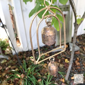 May include: A gold metal wind chime with two bells. The bells are made of a weathered metal and have a rustic look. The wind chime is hanging from a tree branch.