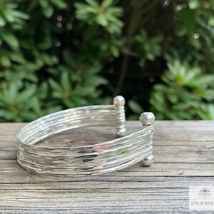 May include: A silver bracelet with a hammered texture and multiple bands. The bracelet has rounded ends.