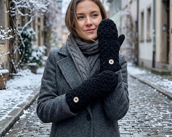 May include: A person wearing black mittens with a white button detail. The mittens are thick and knitted, and the person is wearing a gray coat and scarf. The background shows a cobblestone street and buildings.