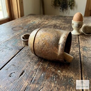 May include: A cylindrical brass bell featuring etched geometric patterns and a floral motif. The bell has a small loop at the top and a wooden clapper extending from the open end. A small logo with the text KVK BOHOFAIR is visible.