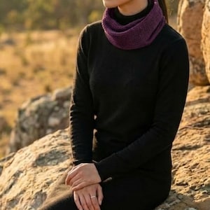 Puede incluir: Una mujer con un vestido negro de manga larga y un calentador de cuello morado est&aacute; sentada en una roca. El fondo presenta un paisaje borroso con luz solar dorada. El atuendo es simple y elegante.