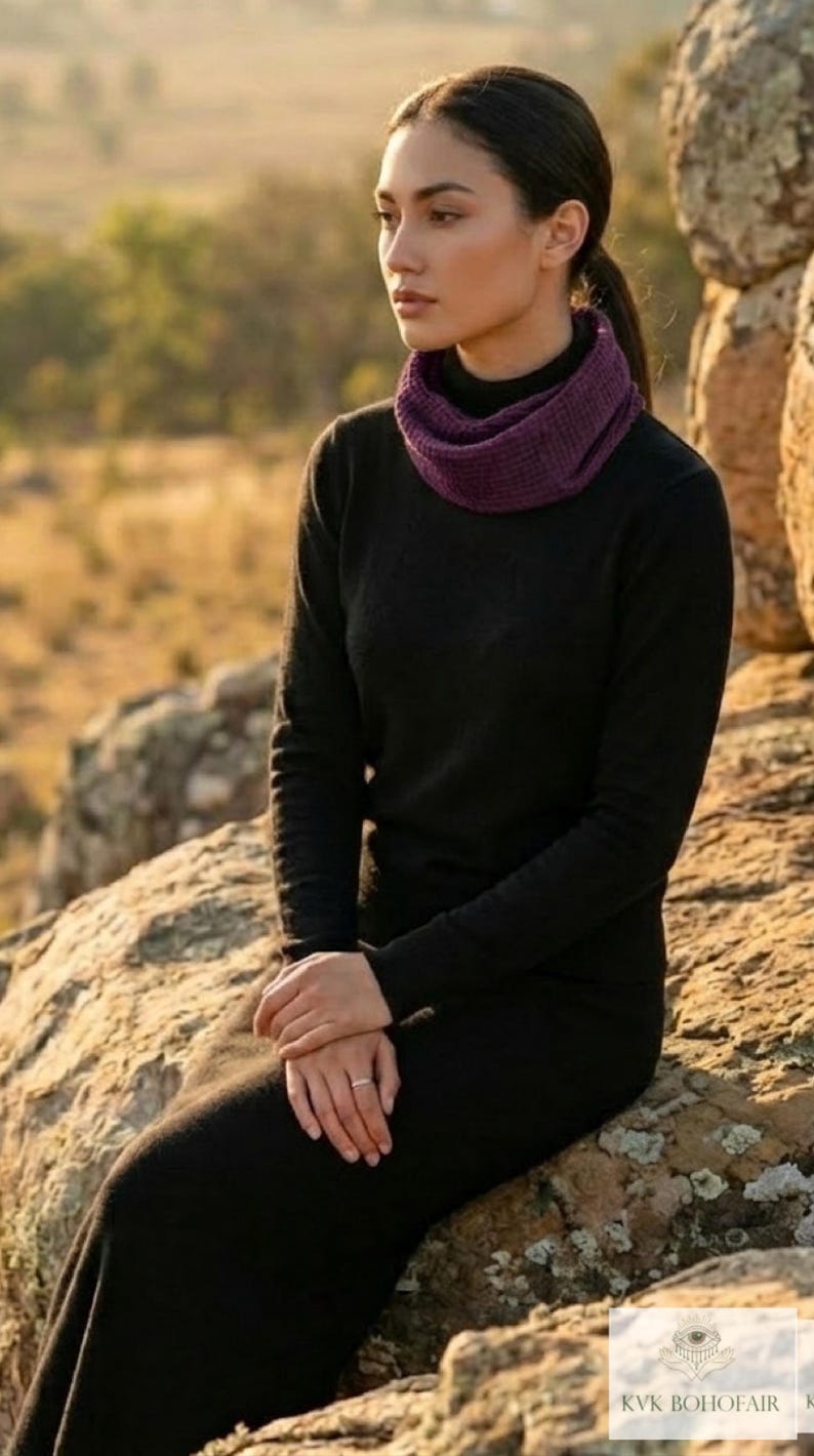 Puede incluir: Una mujer con un vestido negro de manga larga y un calentador de cuello morado est&aacute; sentada en una roca. El fondo presenta un paisaje borroso con luz solar dorada. El atuendo es simple y elegante.