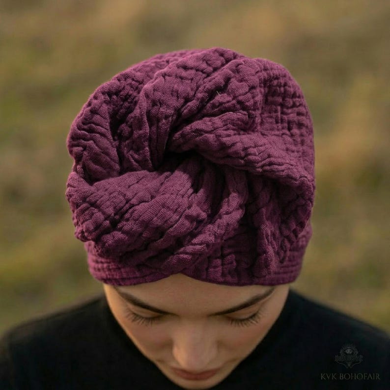 Puede incluir: Un gorro de estilo turbante de color p&uacute;rpura intenso con una apariencia texturizada y acolchada. La tela est&aacute; recogida y torcida en la parte superior, creando un cubrecabezas elegante. El gorro es de color liso y parece estar hecho de un material suave.
