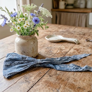 May include: A rustic wooden table displays a bouquet of wildflowers in a beige ceramic vase, a white ceramic spoon, a vintage key, and a blue patterned headband. The headband is tied in a bow.