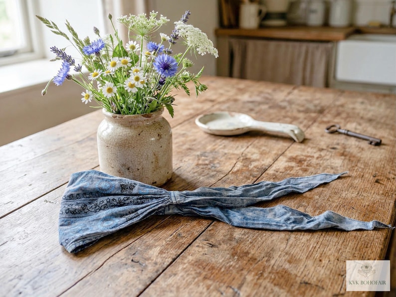 May include: A rustic wooden table displays a bouquet of wildflowers in a beige ceramic vase, a white ceramic spoon, a vintage key, and a blue patterned headband. The headband is tied in a bow.