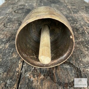 May include: A large, weathered brass bell with a wide, open mouth and a central wooden clapper. The exterior surface features a faint, etched wavy pattern. A small white logo with the text KVK BOHOFAIR is visible in the bottom corner.