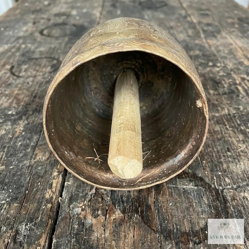 May include: A large, weathered brass bell with a wide, open mouth and a central wooden clapper. The exterior surface features a faint, etched wavy pattern. A small white logo with the text KVK BOHOFAIR is visible in the bottom corner.