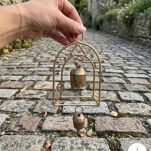 May include: A gold-toned metal arch frame featuring two hanging rustic bells. The structure includes a top circular loop for suspension, a central arch housing the upper bell, and a lower bell suspended from the base of the frame.