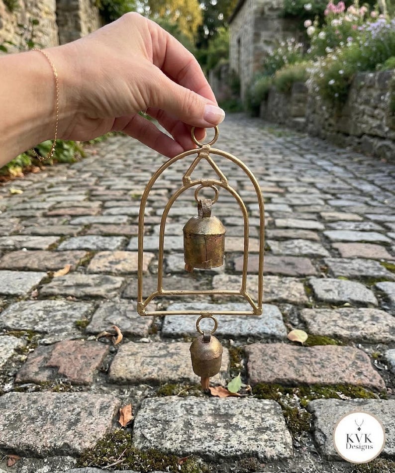 May include: A gold-toned metal arch frame featuring two hanging rustic bells. The structure includes a top circular loop for suspension, a central arch housing the upper bell, and a lower bell suspended from the base of the frame.