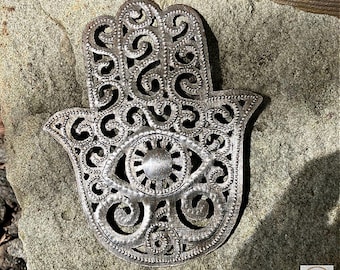 May include: A silver-colored Hamsa hand pendant with an eye design in the center. The openwork design features intricate swirls and patterns. The pendant is displayed on a textured stone surface.