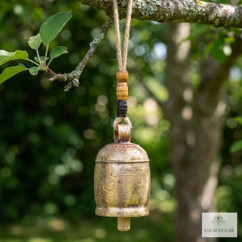 May include: A hanging metal bell with a weathered gold finish and intricate etched patterns. The bell is suspended by a jute twine cord featuring one light wooden bead and one dark wooden bead above the bell's top loop.