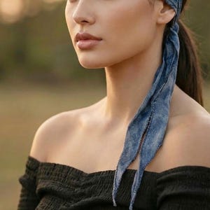 May include: A blue patterned headband with long, flowing ties. The headband is worn with a black off-the-shoulder top. The image is set against a blurred outdoor background, suggesting a bohemian or casual style.