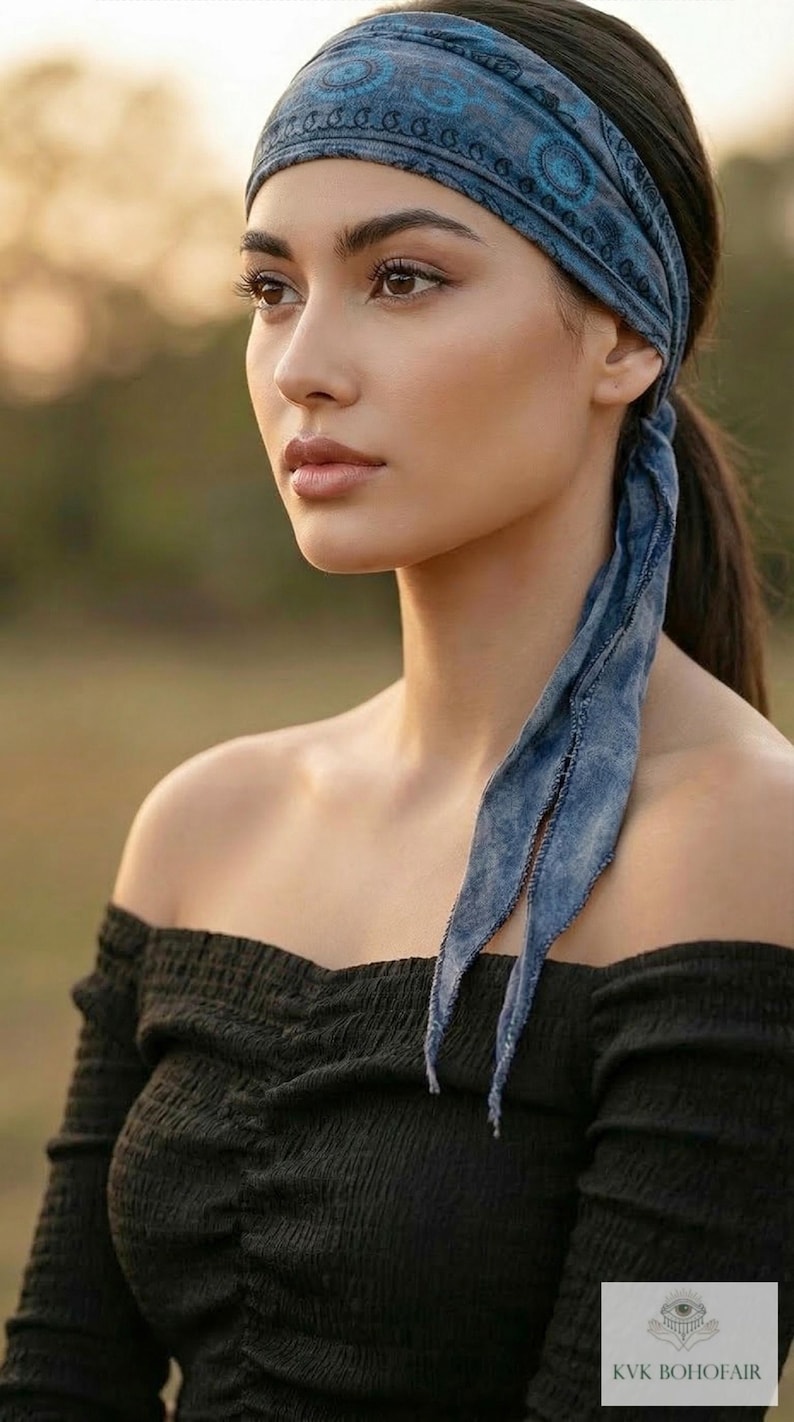 May include: A blue patterned headband with long, flowing ties. The headband is worn with a black off-the-shoulder top. The image is set against a blurred outdoor background, suggesting a bohemian or casual style.