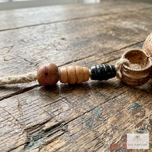 May include: A decorative item featuring a jute cord strung with three distinct beads: a round brown bead with etched markings, a ribbed light brown wooden bead, and a cylindrical black bead, ending in a textured metallic ring.