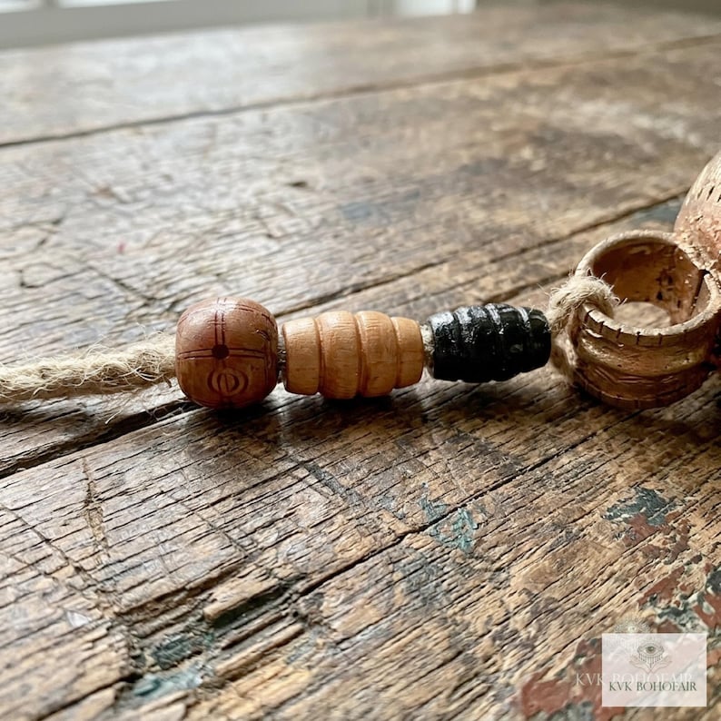 May include: A decorative item featuring a jute cord strung with three distinct beads: a round brown bead with etched markings, a ribbed light brown wooden bead, and a cylindrical black bead, ending in a textured metallic ring.