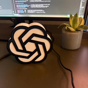 May include: A three-dimensional, circular object featuring a black, interconnected geometric pattern layered over a white base. The design consists of six interlocking loops forming a central hexagon, connected to a black power cord.