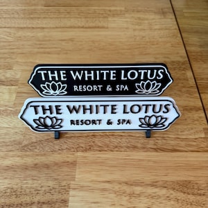 May include: Two signs with the text "THE WHITE LOTUS RESORT & SPA" and a lotus flower graphic. One sign is black with white text and the other is white with black text. Both signs have a decorative arrow shape.