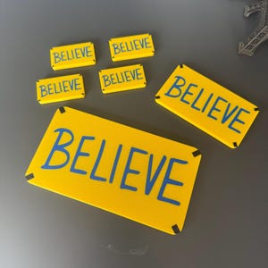 May include: Six yellow signs with black borders and blue lettering that say "BELIEVE". The signs are of various sizes and are arranged on a gray surface.