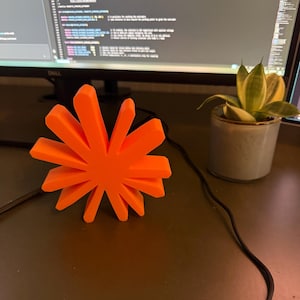 May include: A bright orange, star-shaped object with twelve radiating, flat-edged points. The object has a solid, matte finish and a symmetrical, geometric design, standing upright as a decorative piece on a flat surface.