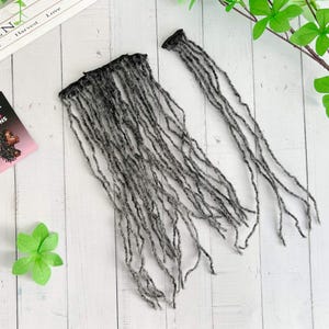 Op de afbeelding: Twee bundels grijze dreadlock-extensions worden getoond op een wit houten oppervlak. Eén bundel is een volledige weft, de andere is een enkele streng. Een kleine kaart met de tekst "Premium Loc Designs" en het profiel van een vrouw is zichtbaar.