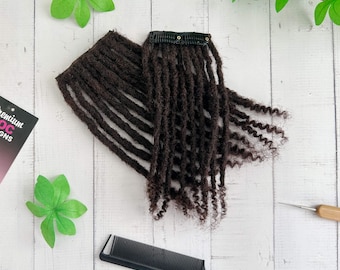 May include: Dark brown dreadlock extensions with a black clip, a black comb, and a crochet hook are displayed on a white wooden surface. Green decorative leaves are scattered around the hair extensions. The extensions are tightly coiled.