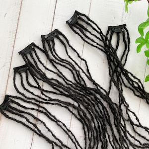 May include: A set of six black dreadlock hair extensions with clips. The extensions are long and straight, and they are arranged in a fan shape.