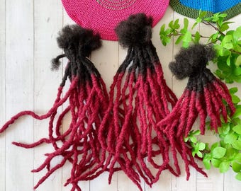 May include: Three sets of synthetic dreadlocks with black roots and burgundy tips. The dreadlocks are arranged on a white wooden surface.