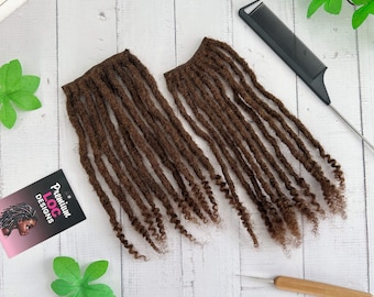 May include: Two bundles of brown dreadlocks are displayed on a white wooden surface, alongside a black comb and a crochet hook. A business card with the text "Premium Loc Designs" is also present. The dreadlocks are neatly arranged, showcasing their texture and color.