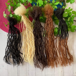 Könnte beinhalten: Fünf Strähnen synthetischer Dreadlocks in verschiedenen Farben: Schwarz, Blond, Braun, Hellbraun und Dunkelbraun. Die Dreadlocks sind in einer Reihe auf einer weißen Holzoberfläche angeordnet.