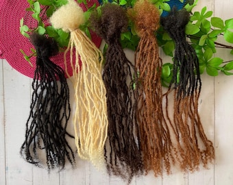 May include: Five strands of synthetic dreadlocks in different colors: black, blonde, brown, light brown, and dark brown. The dreadlocks are arranged in a row on a white wooden surface.