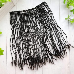 May include: Black dreadlock extensions with multiple clips for easy attachment. The dreadlocks are long and have a natural, textured appearance. The extensions are laid out on a white wooden surface.