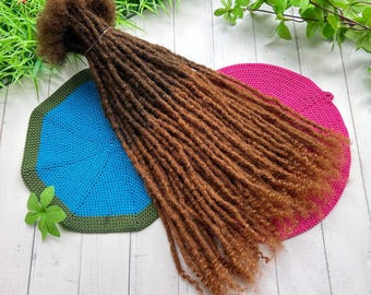 May include: A bundle of brown and auburn dreadlocks, tied at the top, resting on a blue and green crocheted hexagon and a pink crocheted circle. The dreadlocks have a natural, textured appearance, with varying shades of brown.